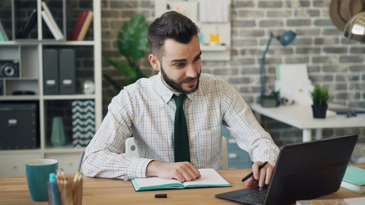 Bărbat la birou lucrând pe laptop, cu un caiet alături