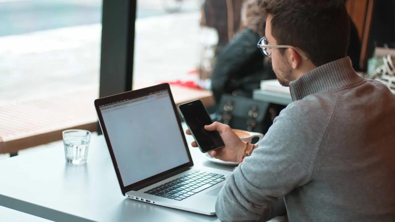Bărbat cu ochelari verificând un email pe telefon, lângă laptop, la o masă