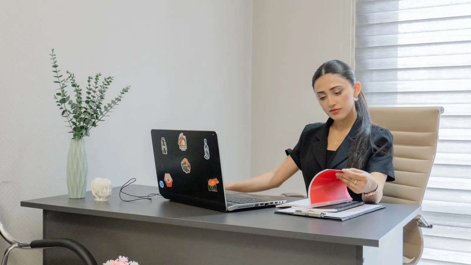 Femeie la birou verificând documente lângă un laptop, într-un birou luminos