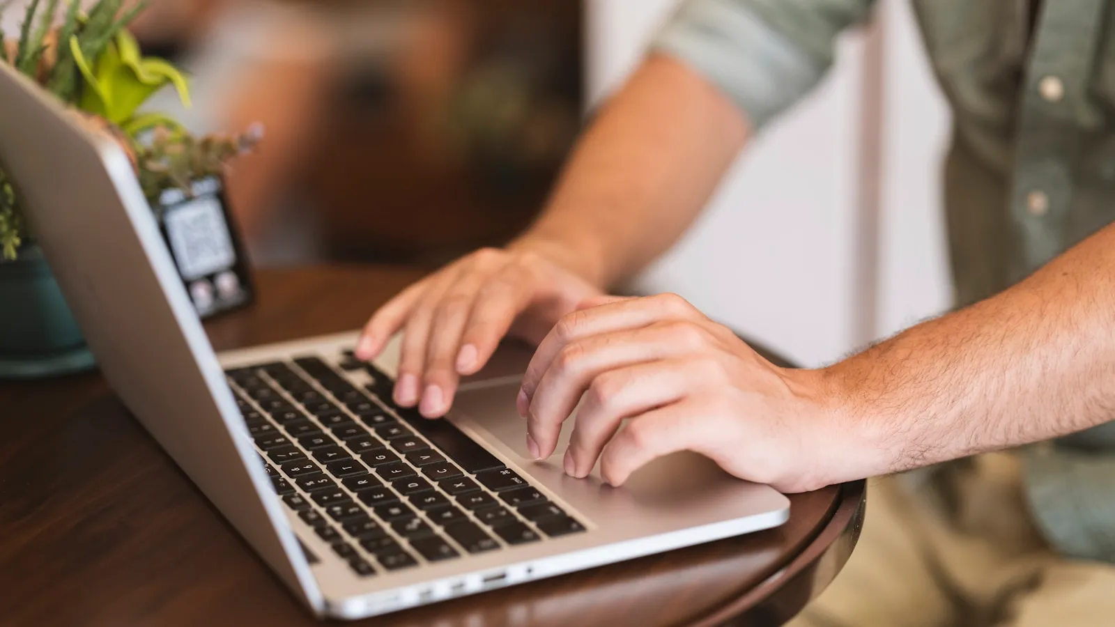 Mâini tastând pe un laptop pe o masă de lemn, lângă o plantă